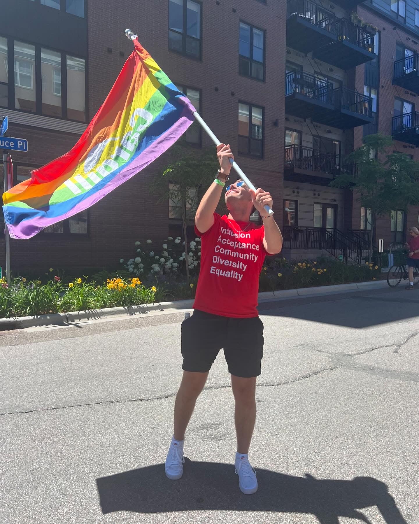 "Man in a red 'Inclusion, Acceptance, Community, Diversity, Equality' t-shirt waving a rainbow Pride flag on a city street."