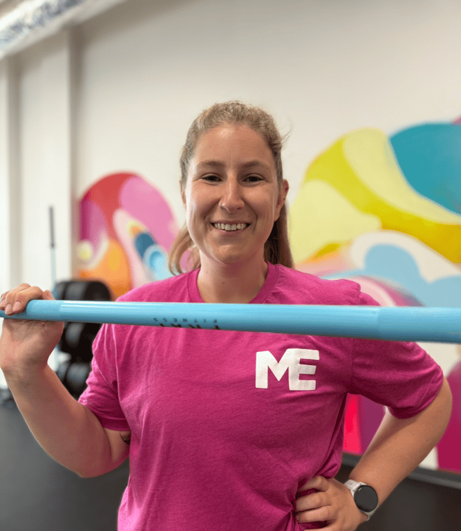 Erin, coach at Studio ME Fitness, smiling in a pink Studio ME shirt holding a bar in front of the colorful studio mural in Northeast Minneapolis.